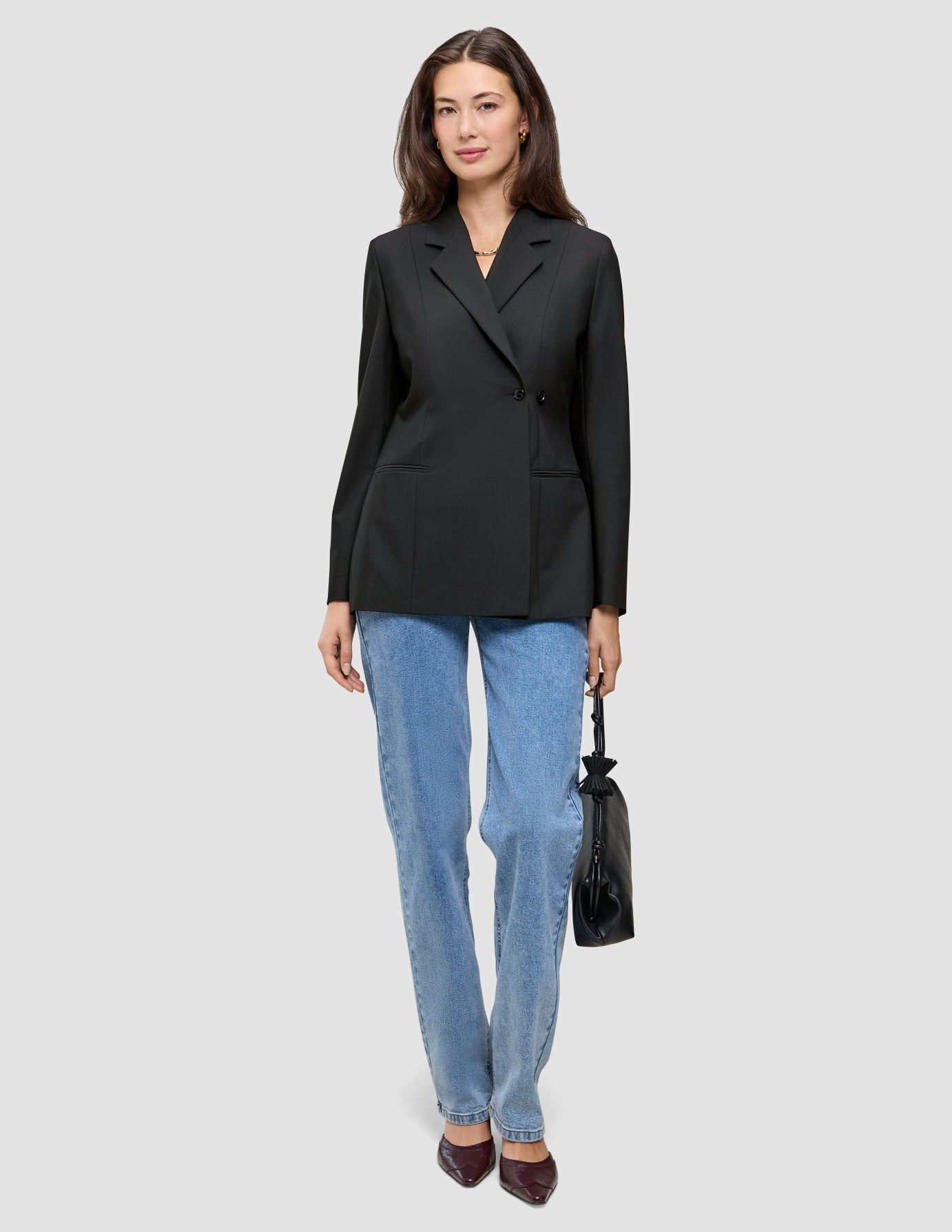 En kvinne har på seg en svart Women’s Avira Blazer Black, blå jeans og mørke høyhælte sko, mens hun holder en liten svart håndveske.